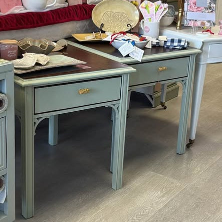 A light sage green vanity desk with a single drawer and gold pull handle, shown in a furniture store showroom with other decor items and wooden flooring.