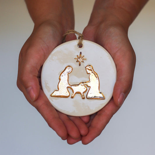 Hands holding a white ornament with gold nativity scene design on a light gray background